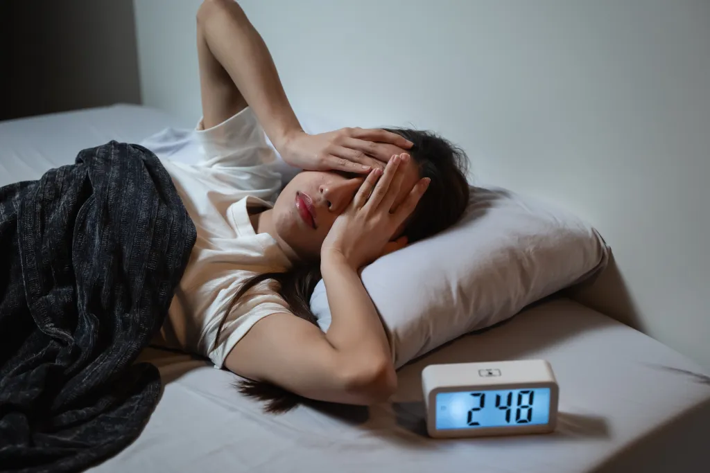 A woman covering her eyes with her hands, lying in bed with a digital clock showing 2:48 next to her, depicting insomnia.