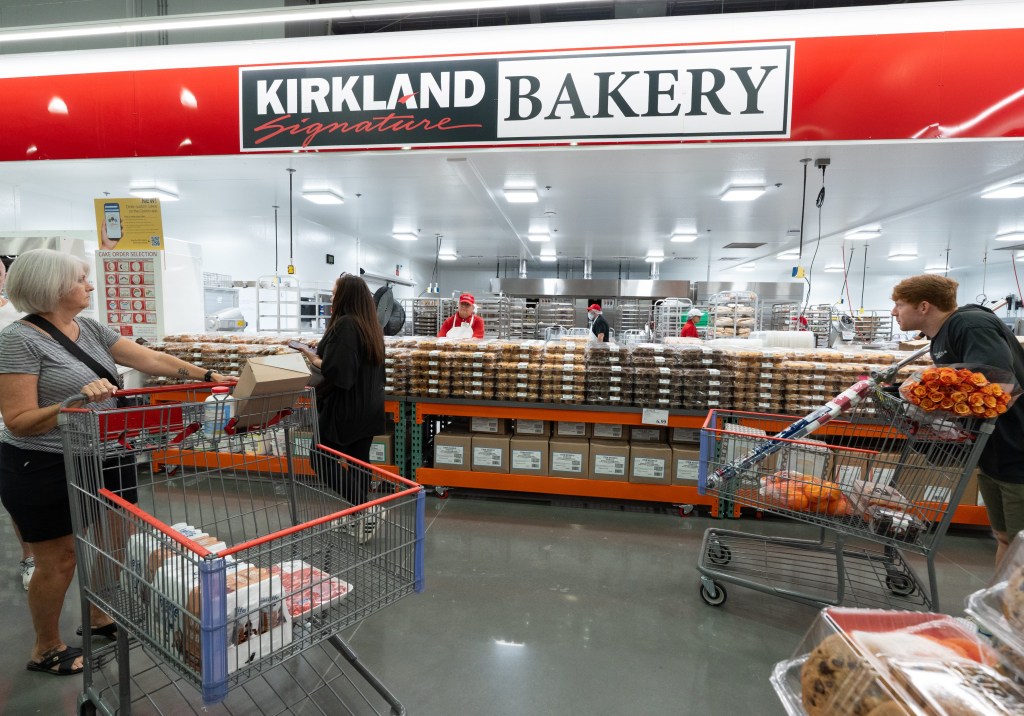A Costco bakery aisle with customers and workers, featuring a large "Kirkland Bakery" sign above shelves stocked with various baked goods.
