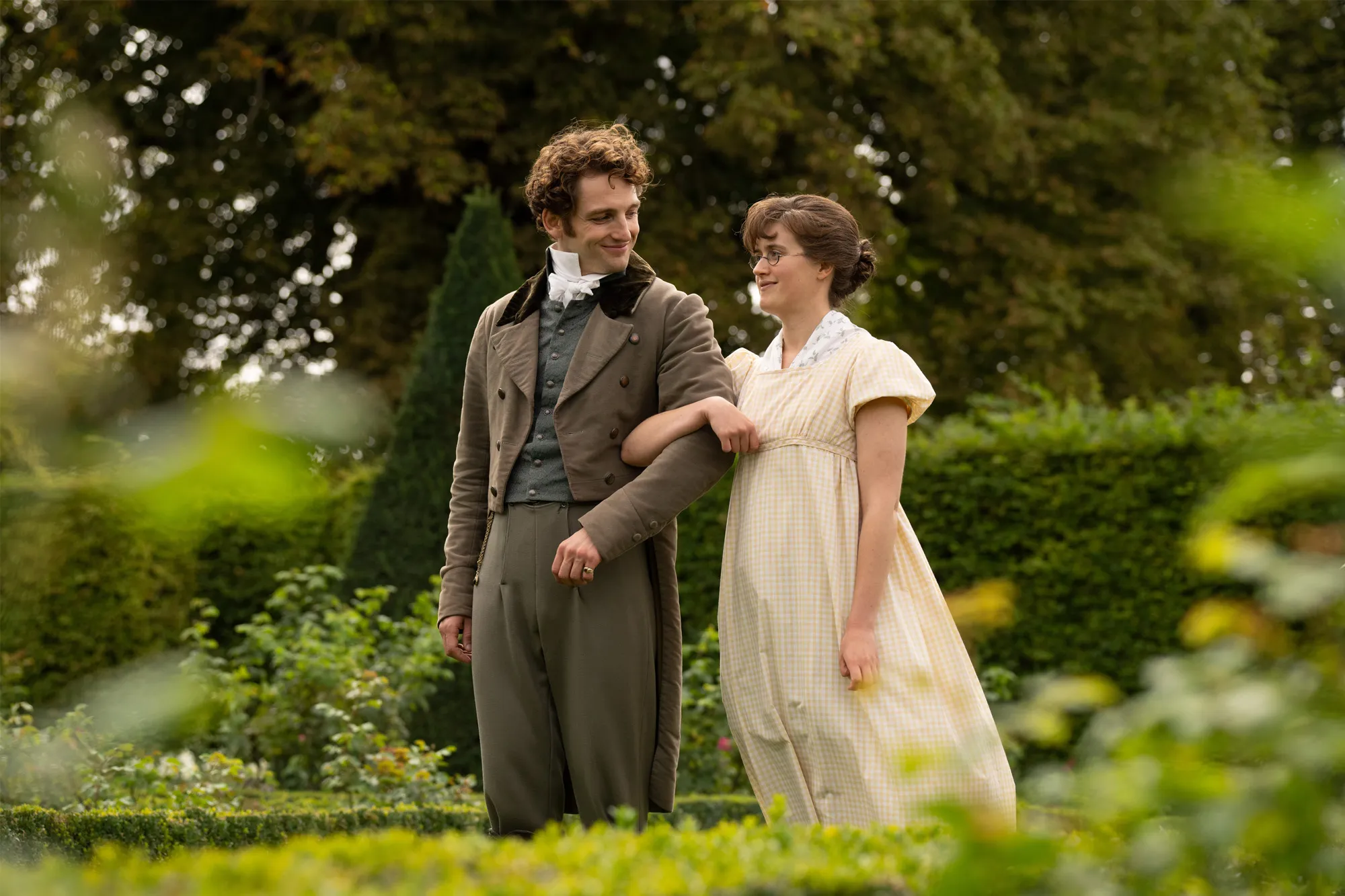 A man in an old-fashioned suit and a woman in a yellow gingham dress arm-in-arm, smiling at each other in a garden.