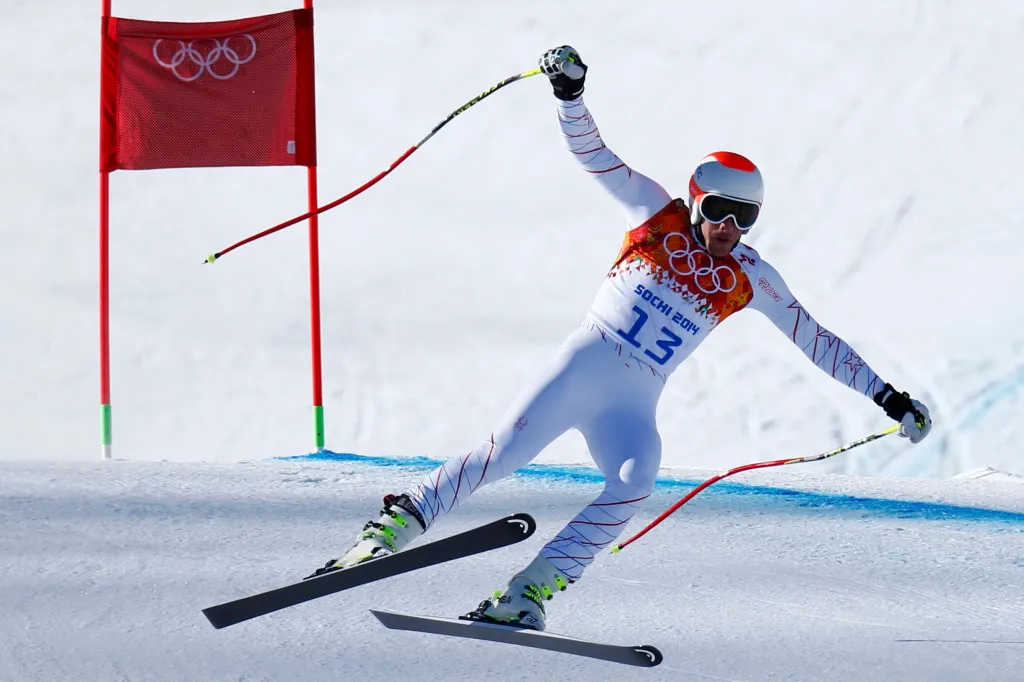 Bode Miller skiing in a white suit with "Sochi 2014" and "13" on it, raising his left arm while going around a red flag with the Olympic rings.