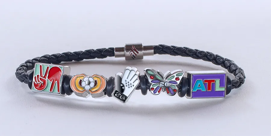A black braided bracelet with five charms: a peace sign, a soccer ball, a baseball glove with "26", a butterfly with "26", and "ATL".