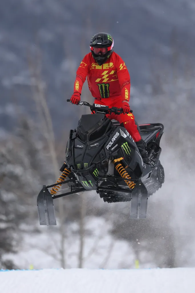 Brett Turcotte in red and yellow racing gear on a black Polaris snowmobile mid-air.