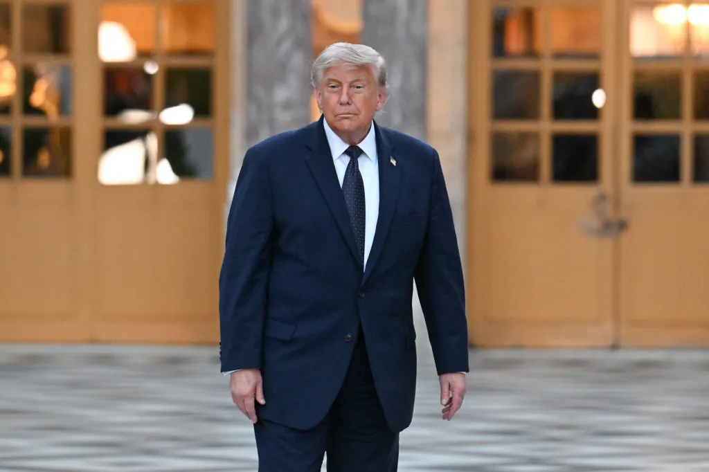 Donald Trump in a navy suit and tie, with a small US flag pin on his lapel, walking past a building.