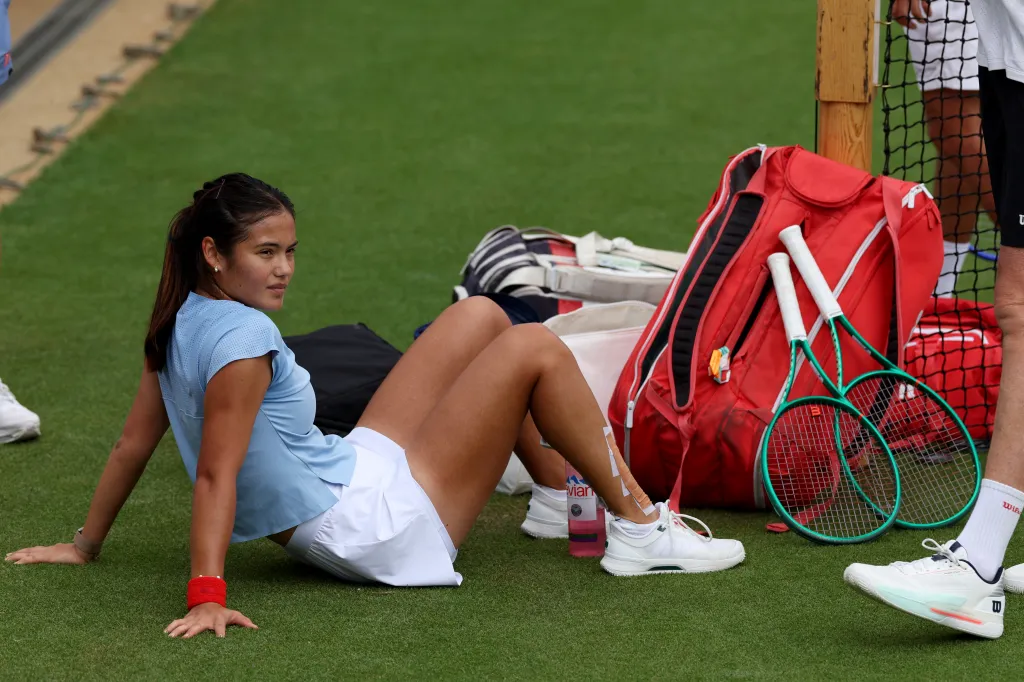 Emma Raducanu rests on the grass with tennis rackets and a bag next to her at Wimbledon.