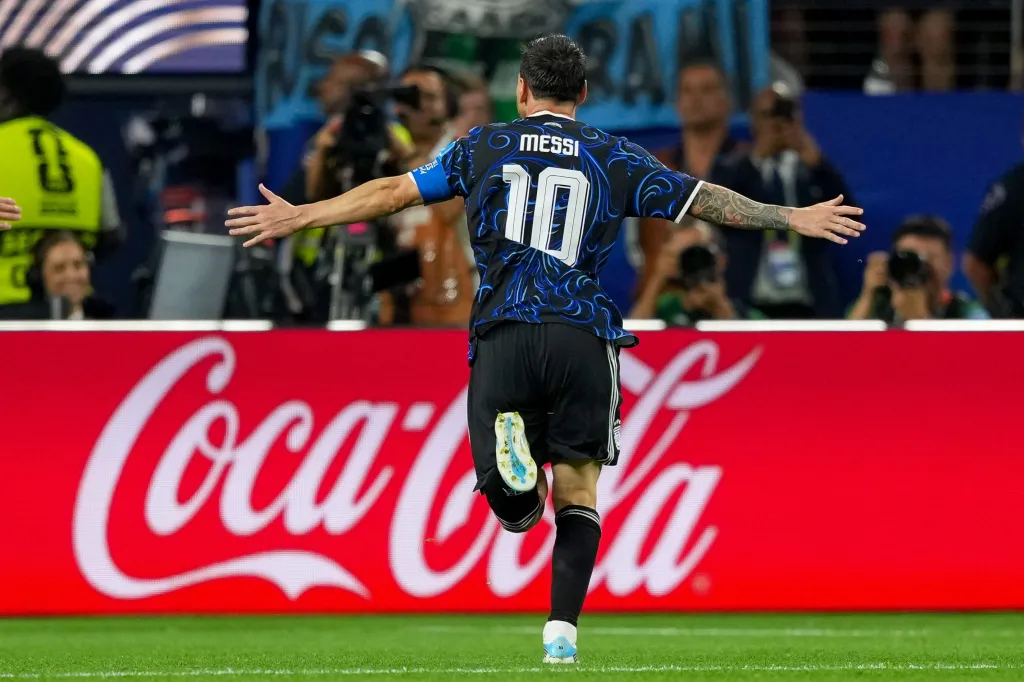 Lionel Messi in a blue and black jersey with number 10, celebrating a goal with arms wide.