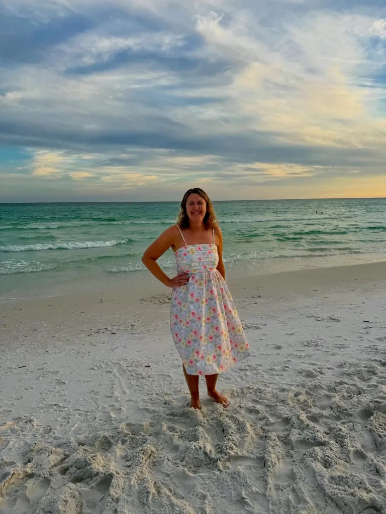 Cheryl Maguire smiling on a beach in a floral dress, with the ocean behind her.