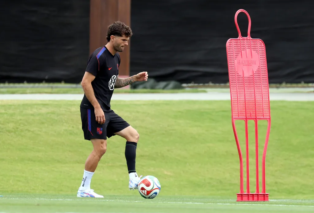 Christian Pulisic of the United States practices with a soccer ball next to a red training dummy on a green field.
