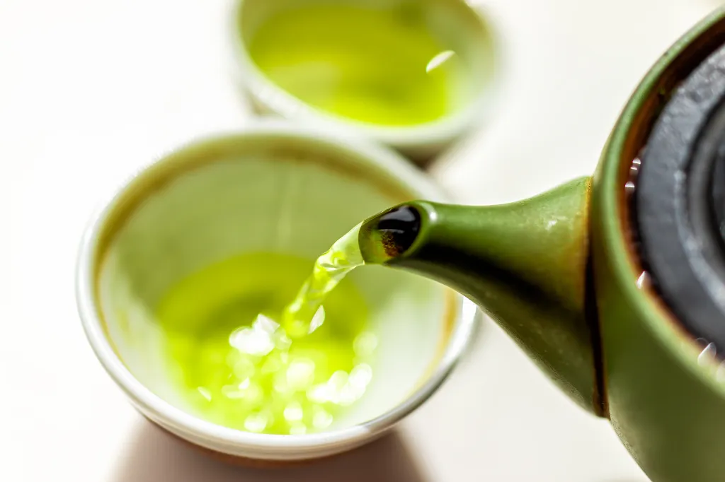 Pouring sencha or genmaicha from a green clay teapot into a ceramic teacup.