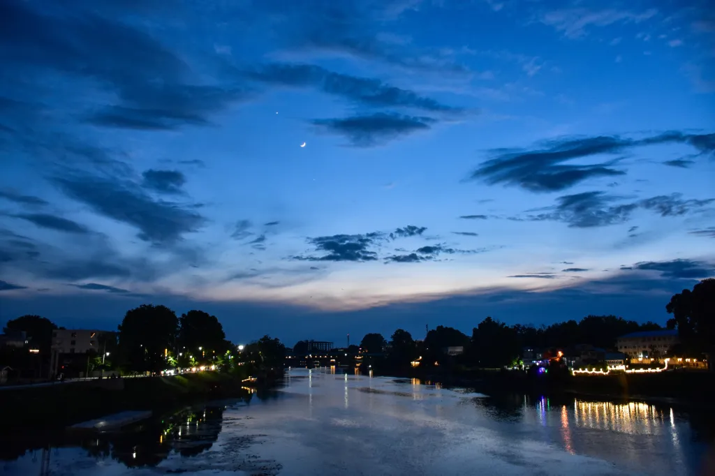 The crescent Moon, Venus, and Jupiter are aligned in the evening sky over a river with city lights along its banks.