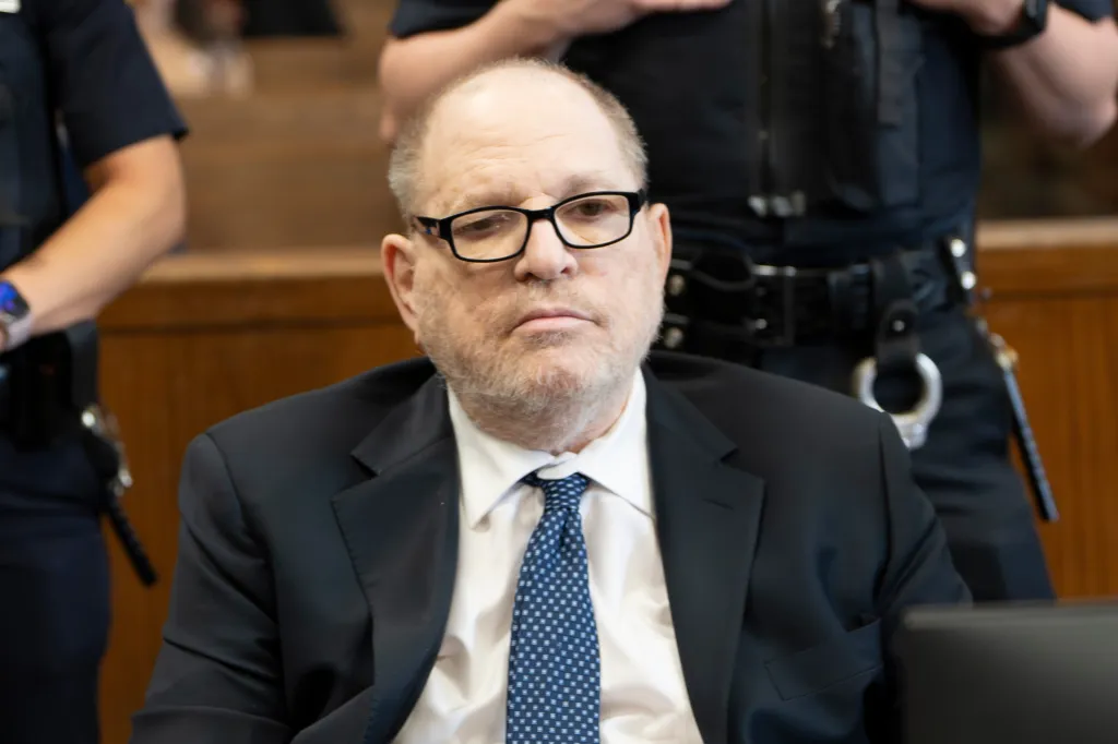 Harvey Weinstein in court, wearing a suit and glasses, with a police officer visible behind him.