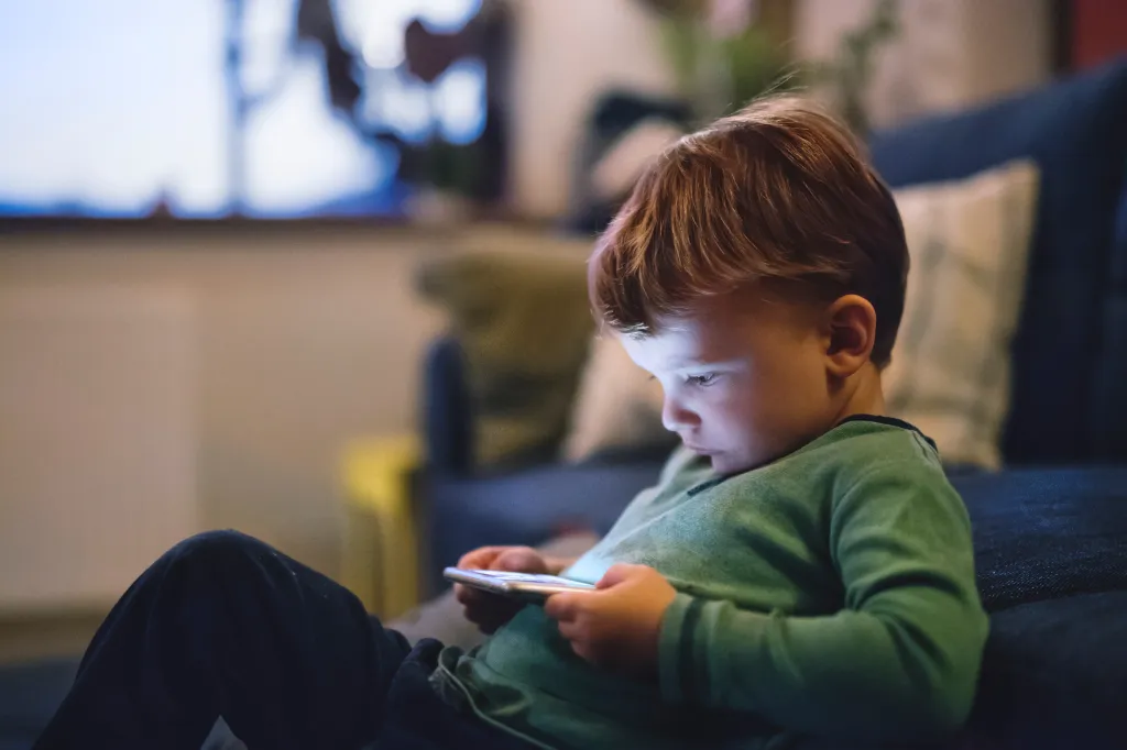 A young boy with reddish-brown hair intently watches a smartphone with blue light reflecting on his face while sitting on a dark couch or bed.