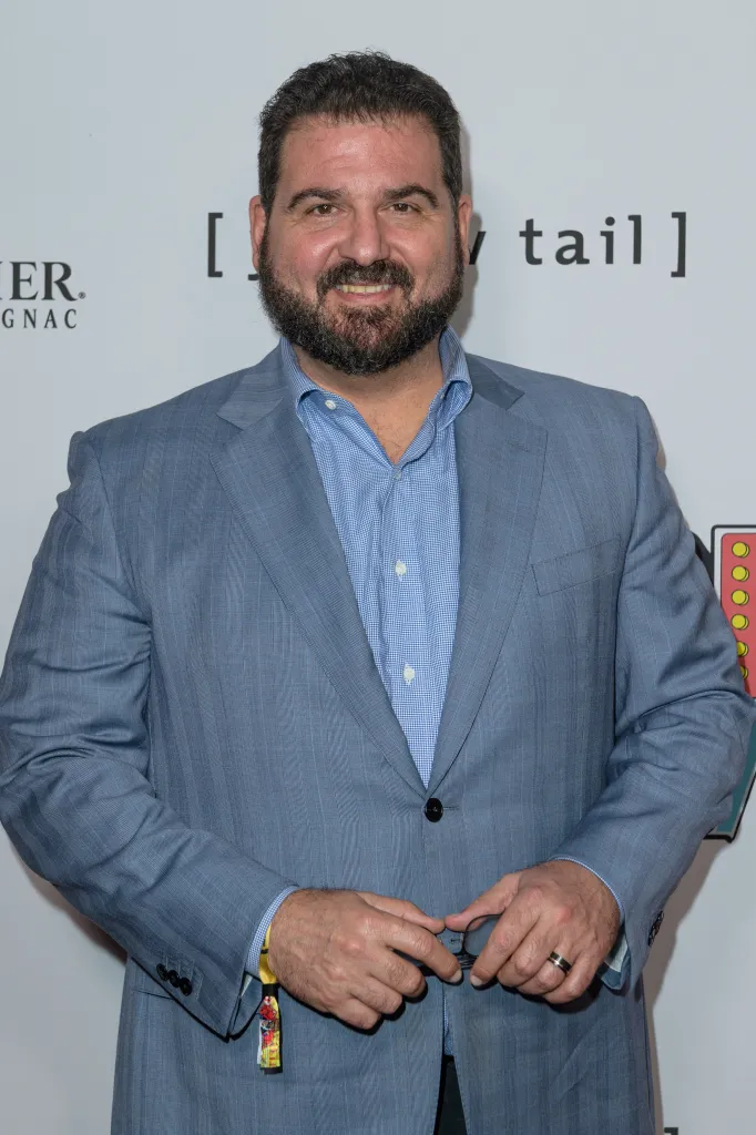 A man with a beard, wearing a light blue suit jacket and a checkered blue shirt, smiles at the camera.