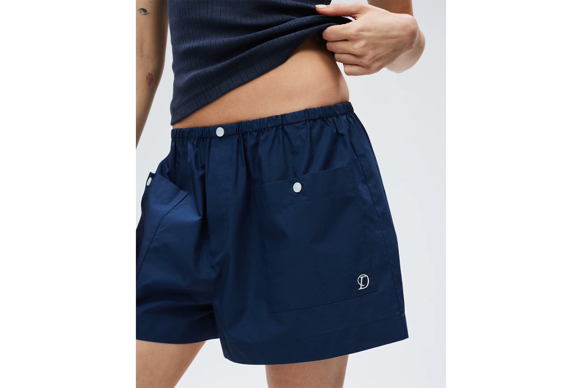 A person wearing navy blue shorts with white buttons and a white embroidered 'D' logo, pulling up a navy blue tank top to reveal their stomach.