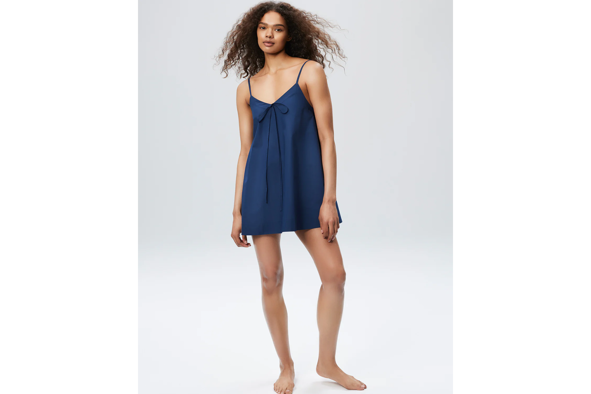 A woman with long curly hair wears a navy blue mini dress with thin straps and a bow tied at the chest.