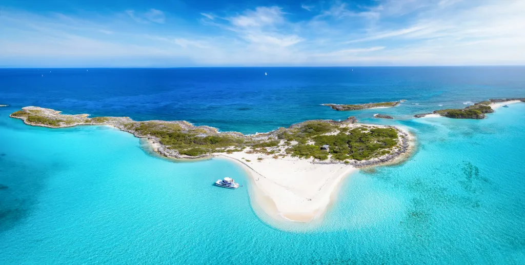 Panoramic aerial view of the Exumas Cays in The Bahamas, showing turquoise waters, white sandbars, and small islands with lush vegetation, with a white boat anchored near a sandy beach.