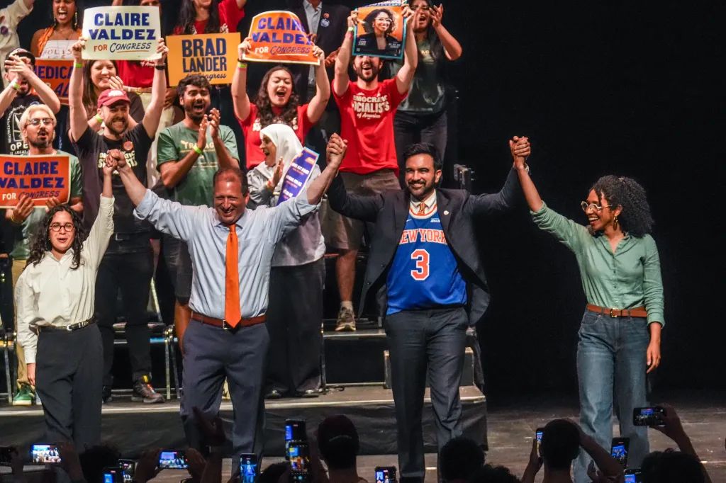 Democratic Congressional candidates Claire Valdez, Brad Lander, and Darializa Avila, along with Zohran Mamdani, holding hands with raised arms on stage at a Get Out The Vote rally.