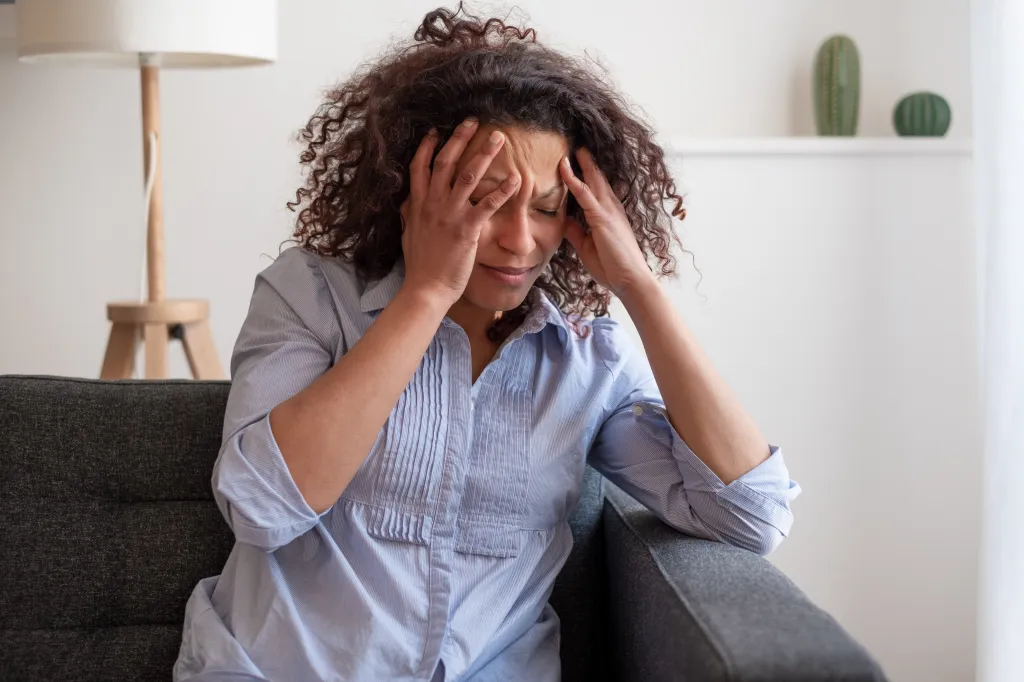 A Black woman sitting on a sofa, eyes closed, massaging her temples due to headache and stress.
