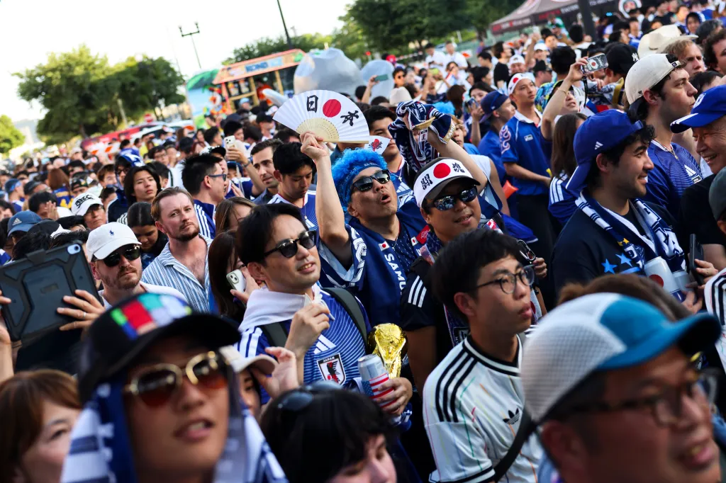 Fans celebrating Japan Day, with one holding a fan with a Japanese flag design and another holding a replica of the World Cup trophy.
