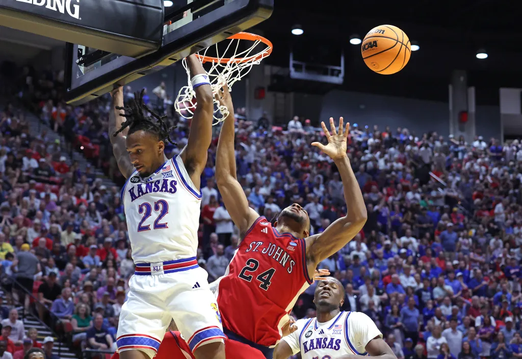 Kansas Jayhawks guard Darryn Peterson #22 hangs on the rim after a dunk, while St. John's Red Storm forward Zuby Ejiofor #24 attempts to block the ball from going in the net, with a Kansas Jayhawks player #0 looking on.
