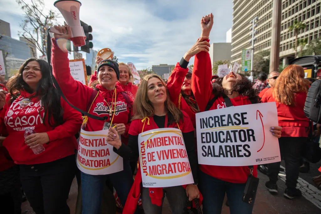 A crowd of people in red shirts at a rally, holding signs that say "Increase Educator Salaries" and "Justicia Ambiental de Vivienda Migratoria."