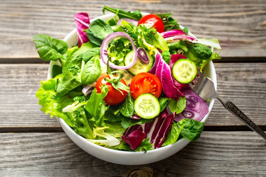 Fresh green salad with tomatoes, cucumbers, and red onion in a white bowl, with a fork, on a wooden background.
