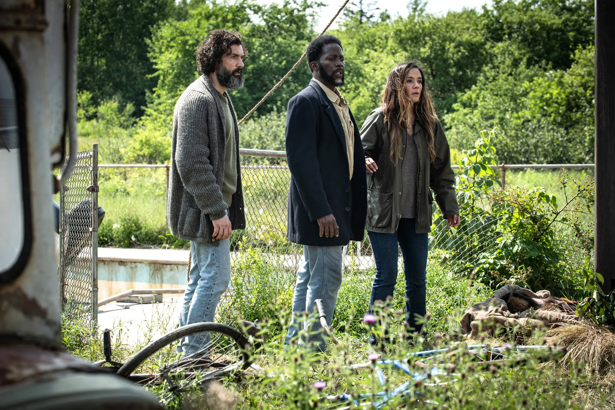 Three people standing outside near a fence and overgrown vegetation.