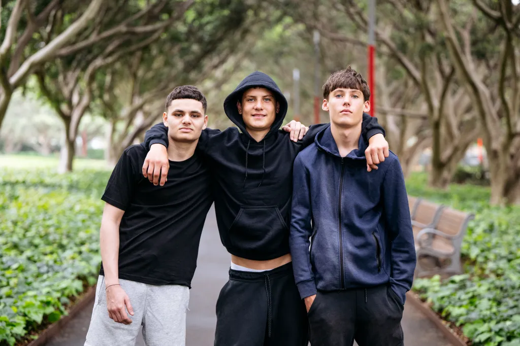 Three teenage boys in athletic wear stand with arms around each other's shoulders.