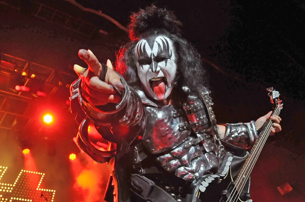 Gene Simmons of Kiss, with face paint and armor, performing on stage with his bass guitar and tongue out.
