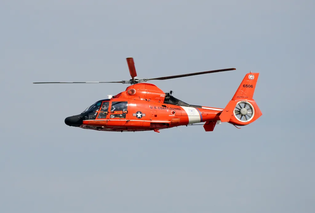 A U.S. Coast Guard helicopter in flight, showing its orange and white livery, with "U.S. COAST GUARD" and the number "6506" visible.