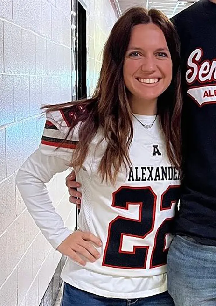 A smiling woman with long brown hair wearing a white "Alexander 22" football jersey and jeans.