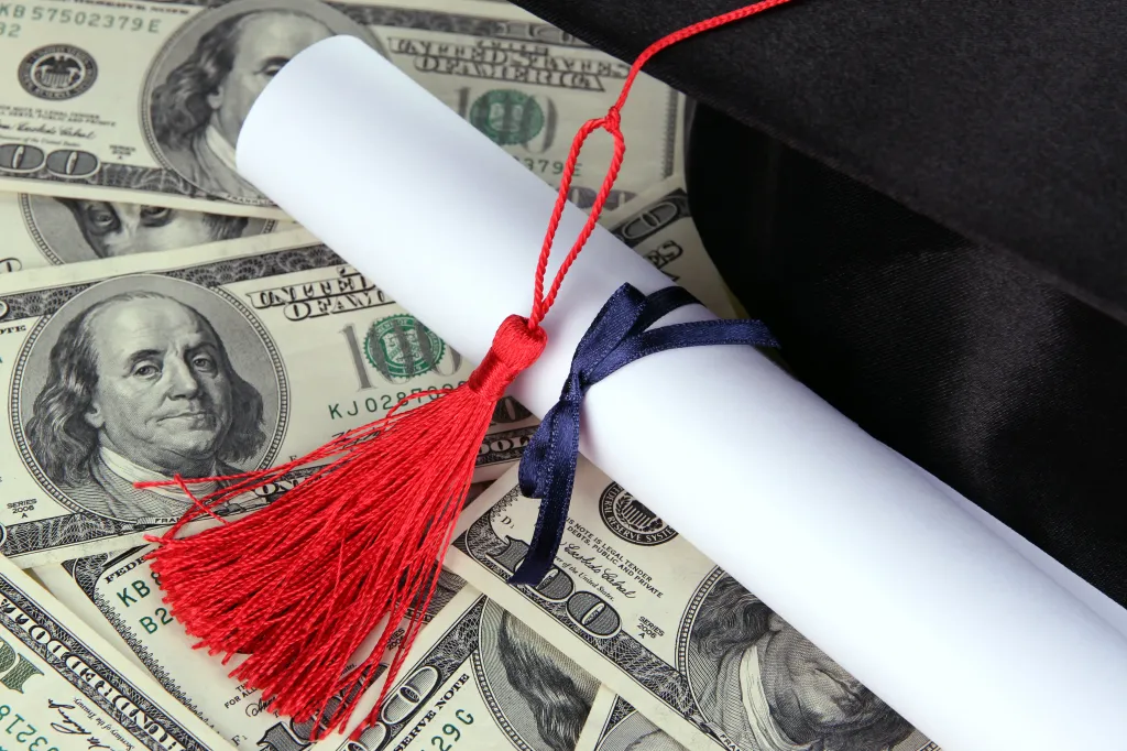 Graduation cap and diploma scroll with a red tassel and blue ribbon, lying on a bed of US hundred-dollar bills.