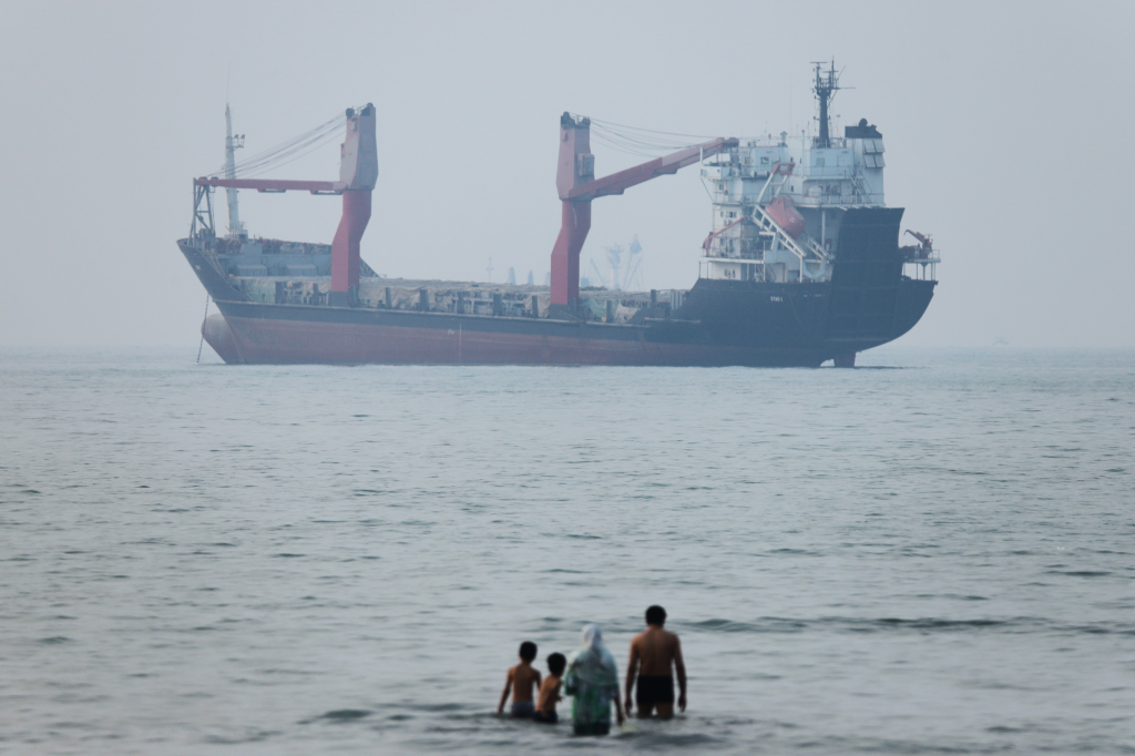 Cargo ship anchored in the Strait of Hormuz as people stand in the shallow water.