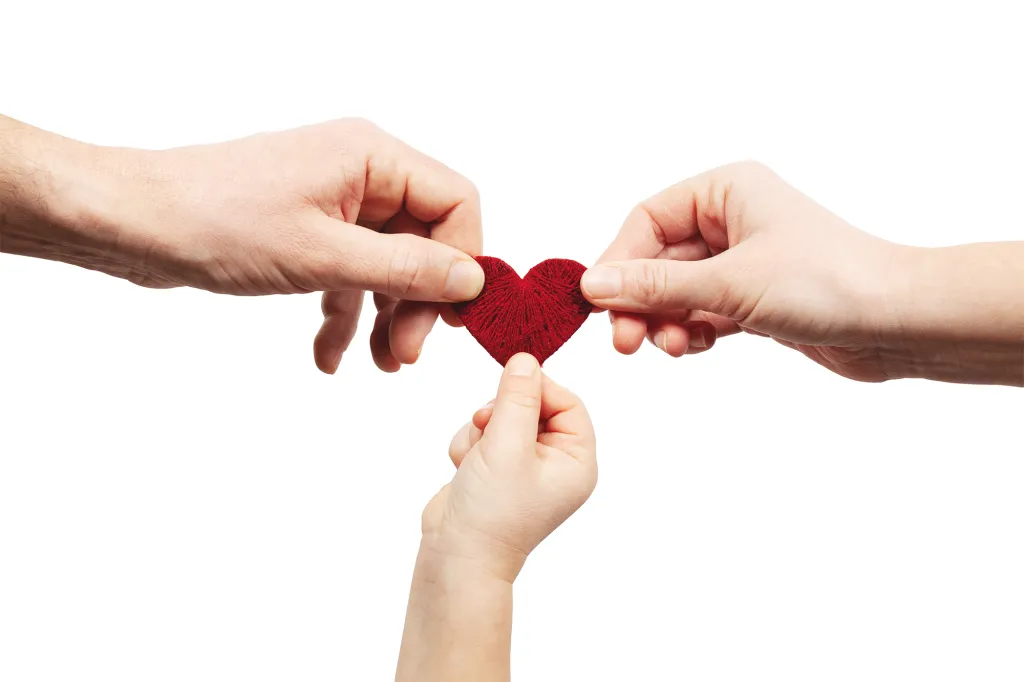 Hands of an adult man, an adult woman, and a child holding a red heart made of yarn.