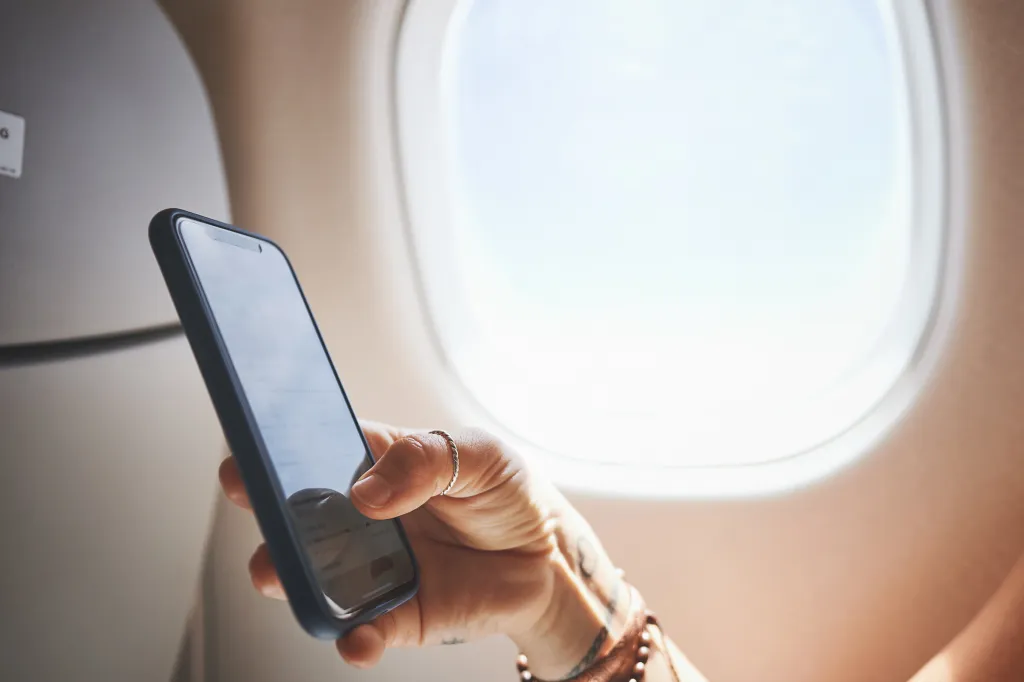A close-up of a person's hand holding a smartphone in an airplane cabin, with a bright window in the background.