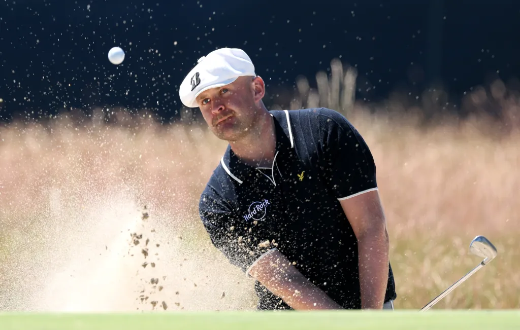 Harry Hall hits a golf shot from a sand bunker, sending sand and the golf ball flying.