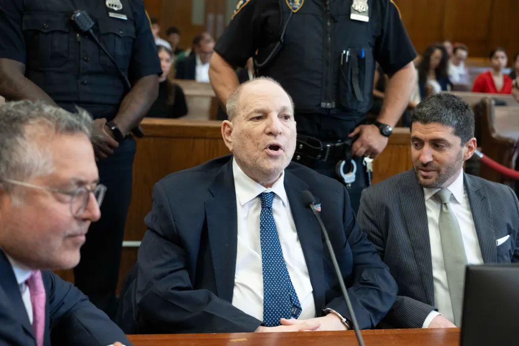 Harvey Weinstein in court, speaking, with two police officers and a lawyer beside him.