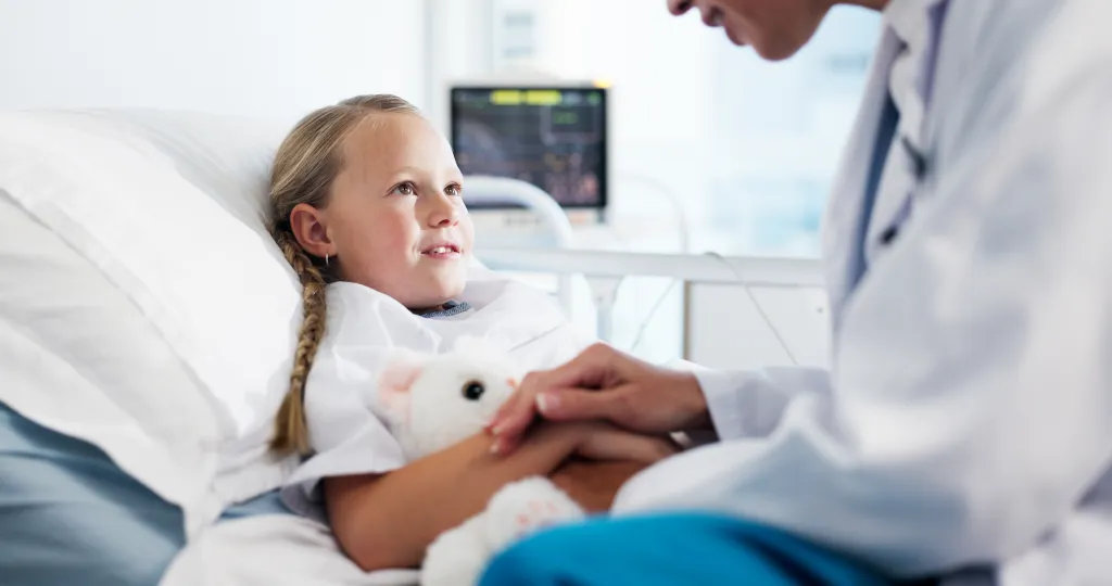 A doctor holding the hand of a child patient with a teddy bear in a hospital bed.
