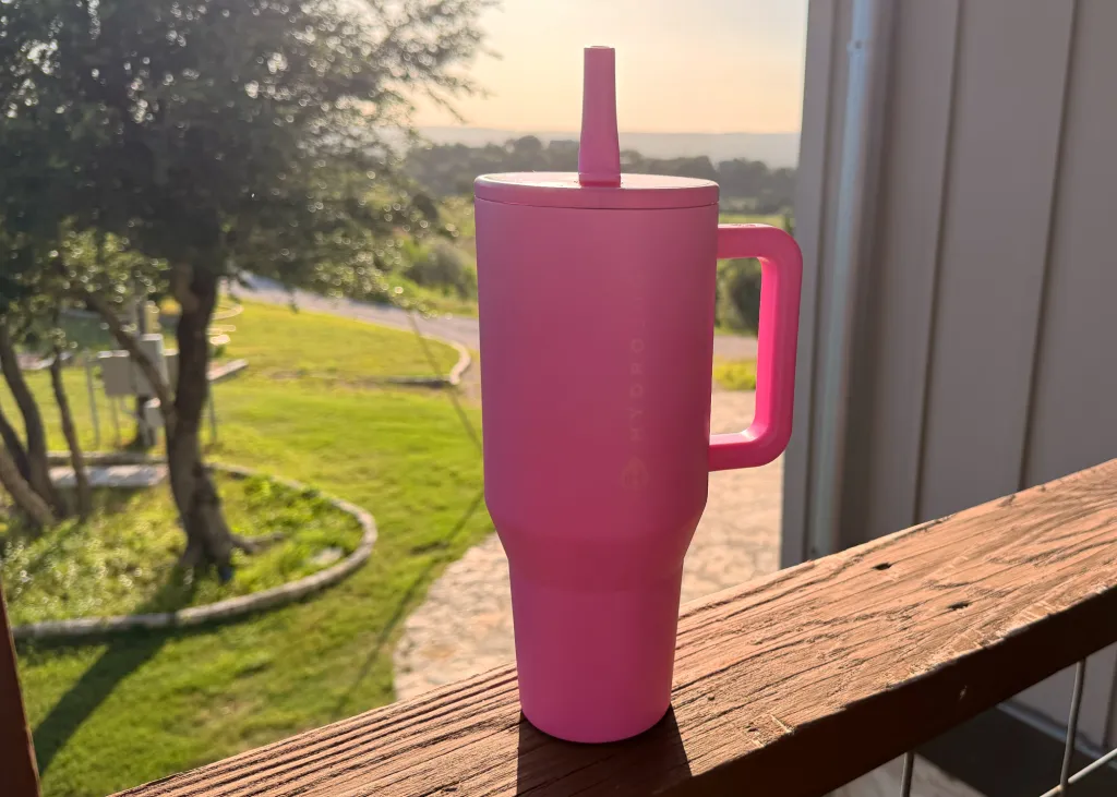 A pink HydroJug brand tumbler with a handle, sitting on a wooden railing.