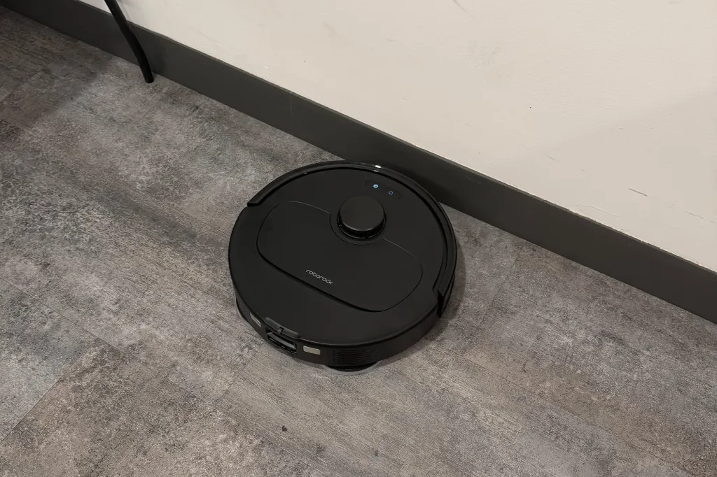 Black Roborock robot vacuum on a dark gray wood-look floor against a white wall.
