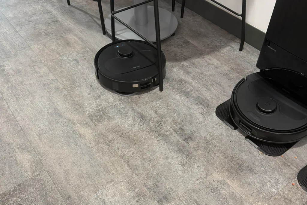 Two black Roborock robot vacuums, one charging against a wall and another under a table.