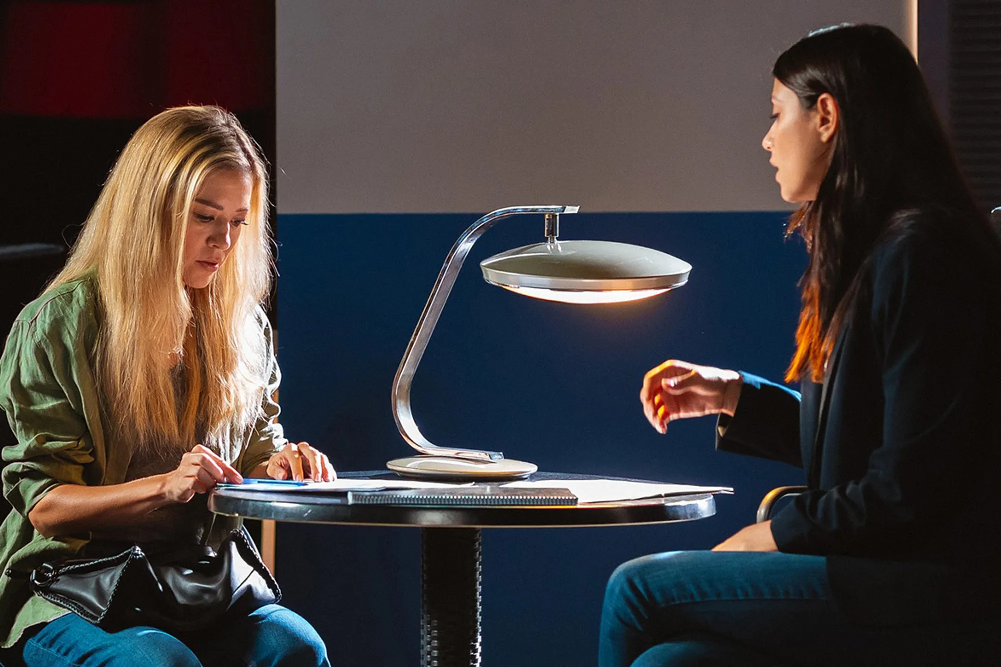 Two women sitting at a table with an illuminated lamp, one looking down and writing, the other looking on.