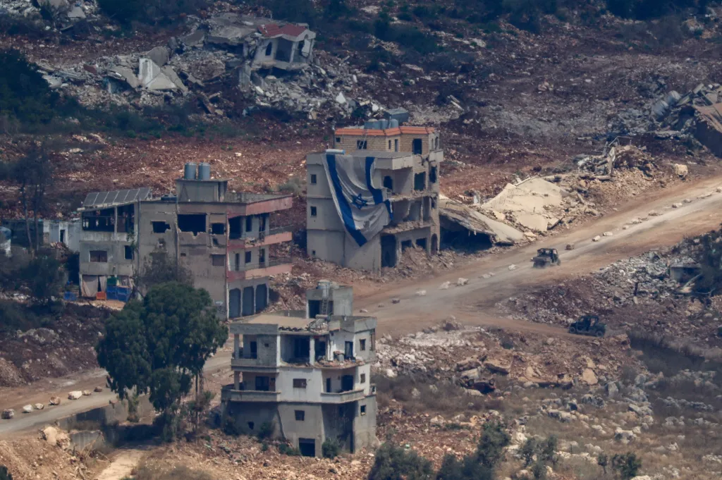 Israeli military vehicles driving on a dirt road near damaged buildings, one draped with an Israeli flag, in Lebanon as seen from northern Israel.