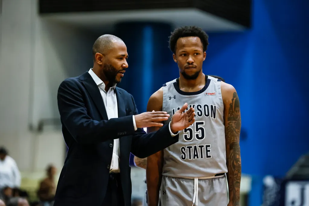 A man in a suit talking to a basketball player wearing a "Jackson State 55" jersey.