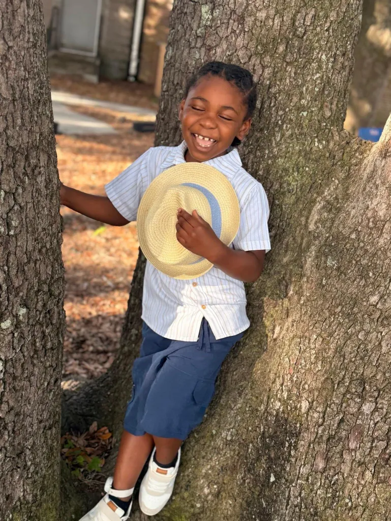 JayCeon Gates smiling while holding a hat and leaning against a tree.