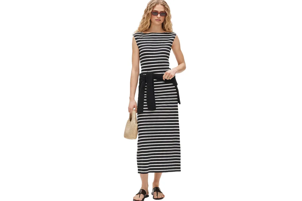 A woman in a black and white striped boatneck maxi dress with a black sweater tied around the waist, black flip-flops, and a small straw handbag.