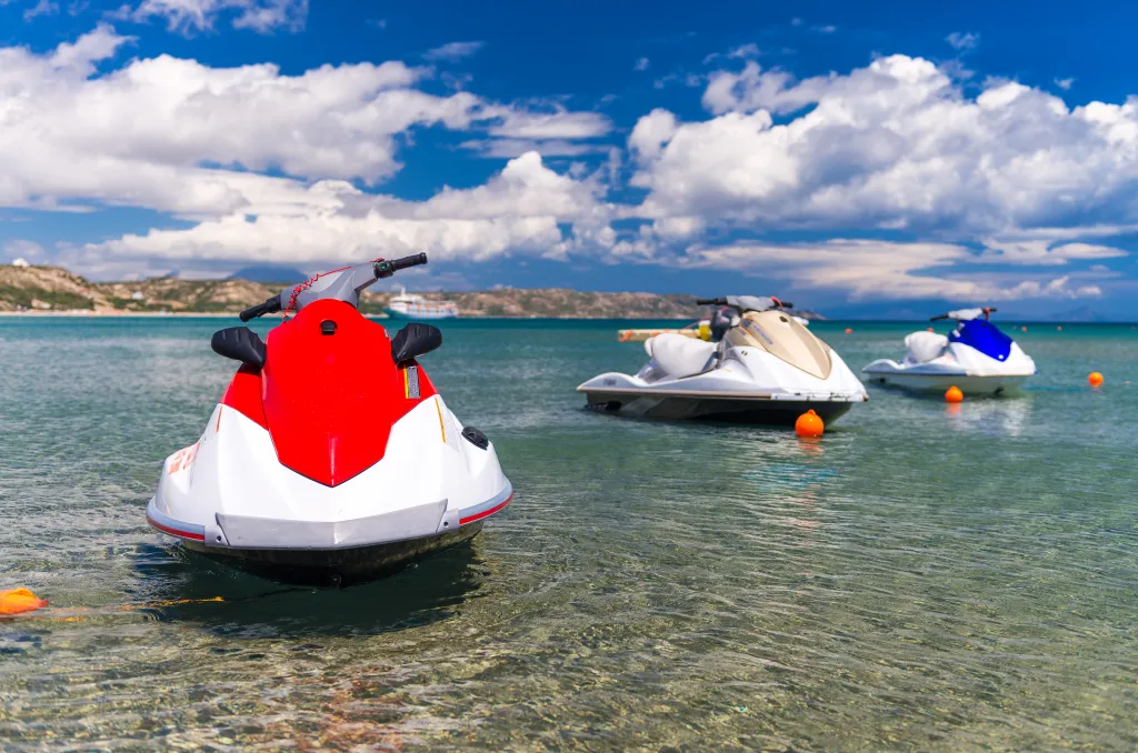 Three jet skis, one red and white, one gold and white, and one blue and white, anchored in clear shallow water near a beach under a blue sky with white clouds.