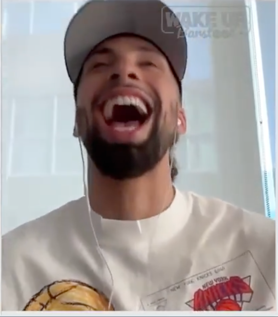 A man with a beard, wearing a baseball cap and a white t-shirt, laughs loudly with his mouth wide open.