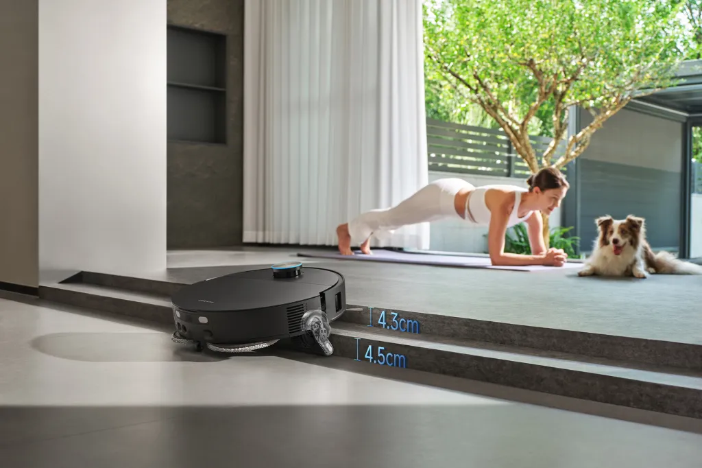 A robot vacuum with an extended brush next to two steps, measuring 4.3 cm and 4.5 cm, while a woman planks on a yoga mat and a dog rests in the background.