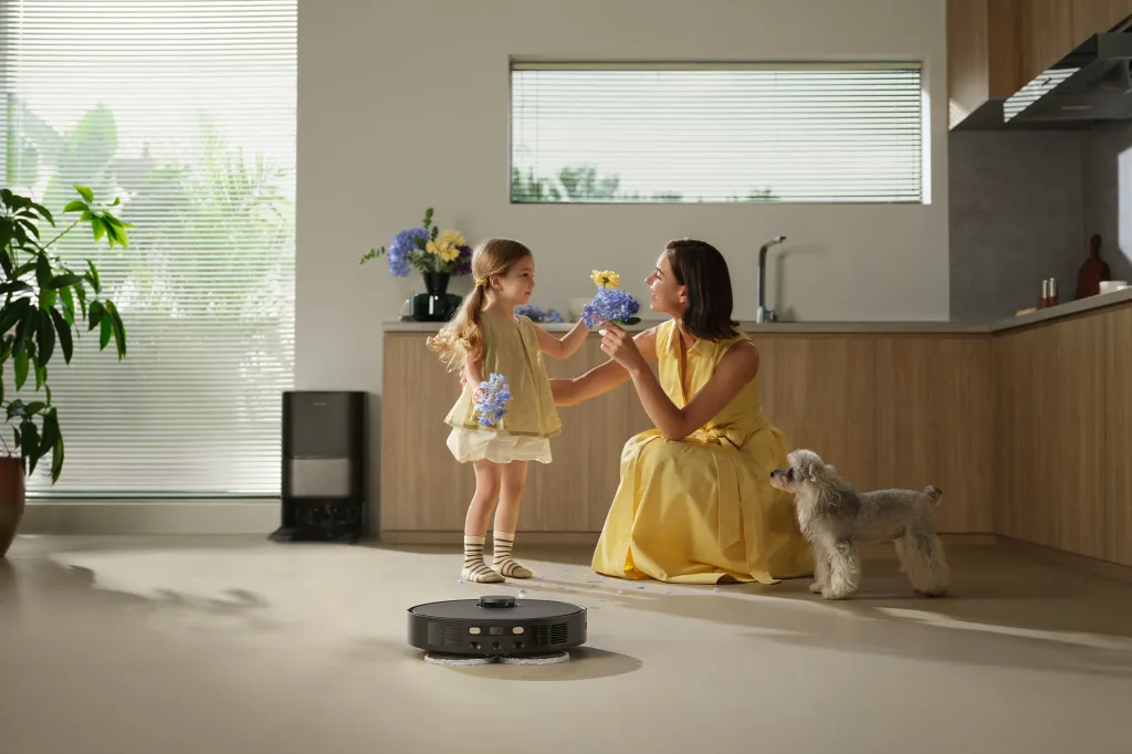 A robotic vacuum cleaner cleaning a kitchen floor while a woman and child play with flowers, and a dog stands nearby.