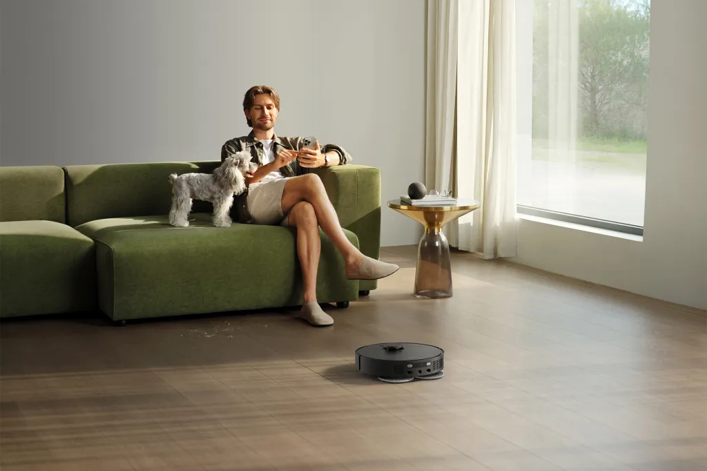 A man sitting on a green couch with a dog next to him while a robot vacuum cleans the floor.